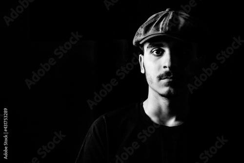 Black and white portrait of a young guy with a hat on black background.