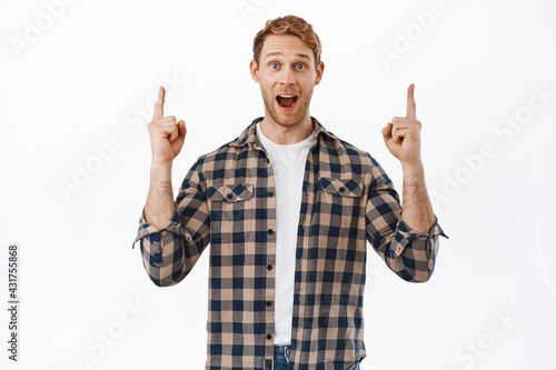 Image of happy man making announcement, redhead guy gasping, saying wow and pointing fingers up, standing impressed with promo deal against white background