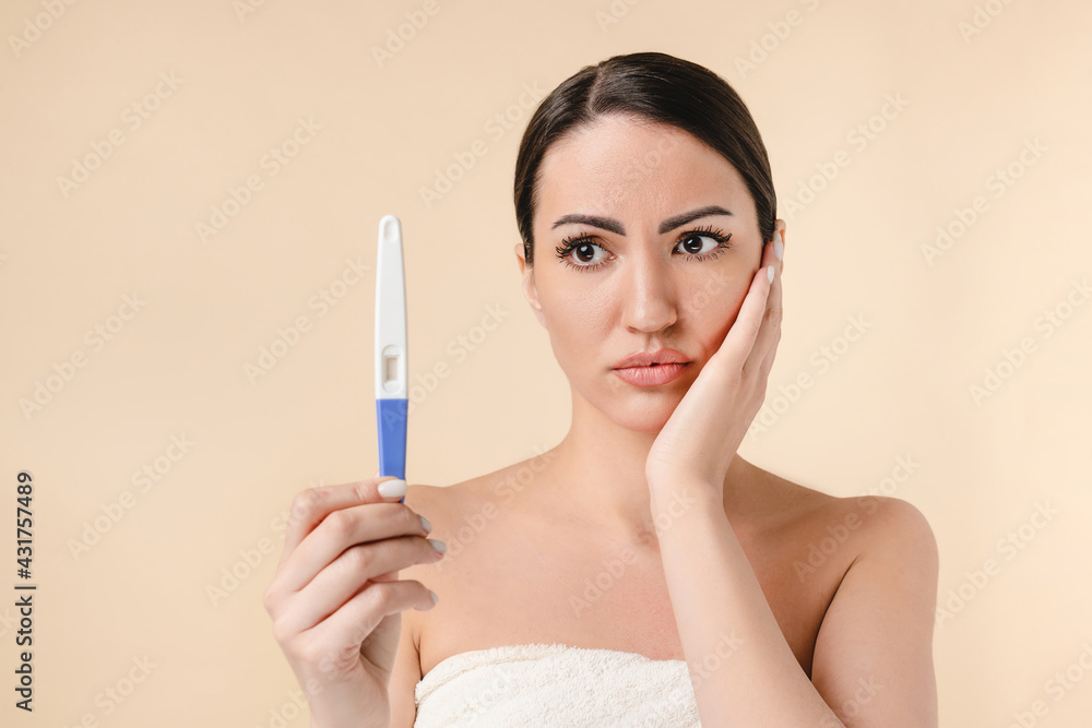 Worried caucasian young pretty woman in bath spa towel holding sadly with fear positive pregnancy test isolated over beige background. Fertility, motherhood, natality concept. Expecting a baby