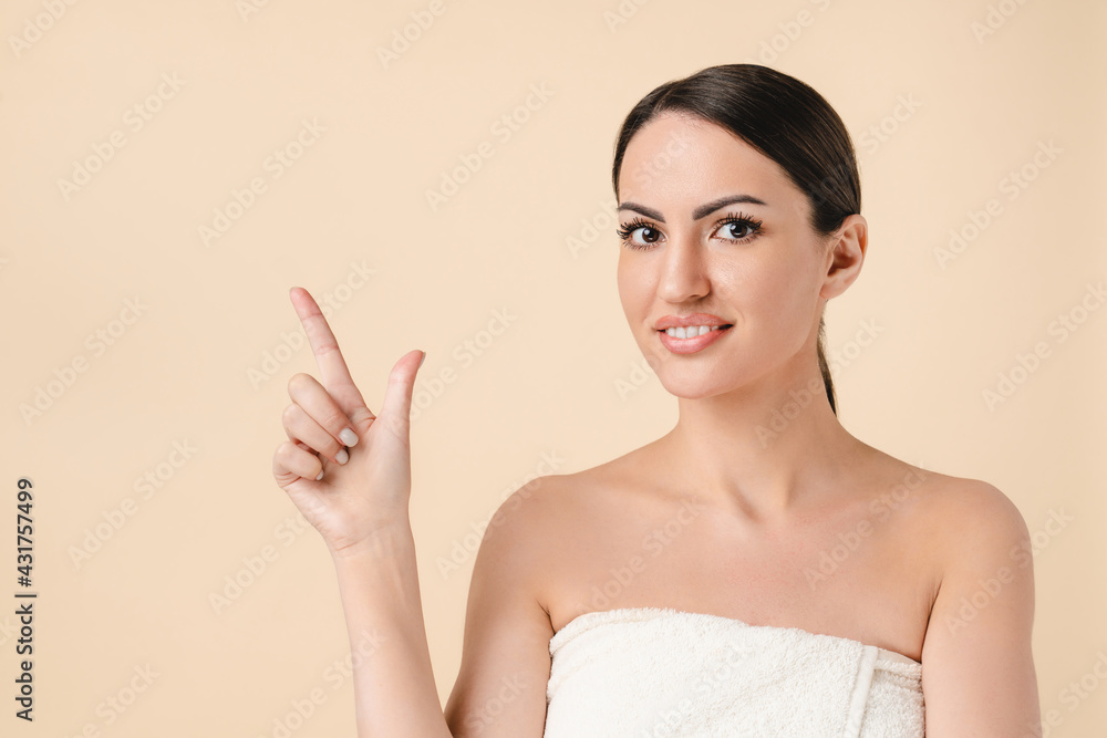 Caucasian beautiful young woman in bath spa towel pointing at copy space isolated over beige background. Pretty young white woman ready for beautification and spa, cosmetology, make up concept. Caucasian beautiful young woman in bath spa towel pointing at copy space isolated over beige background. Pretty young white woman ready for beautification and spa, cosmetology, make up concept.