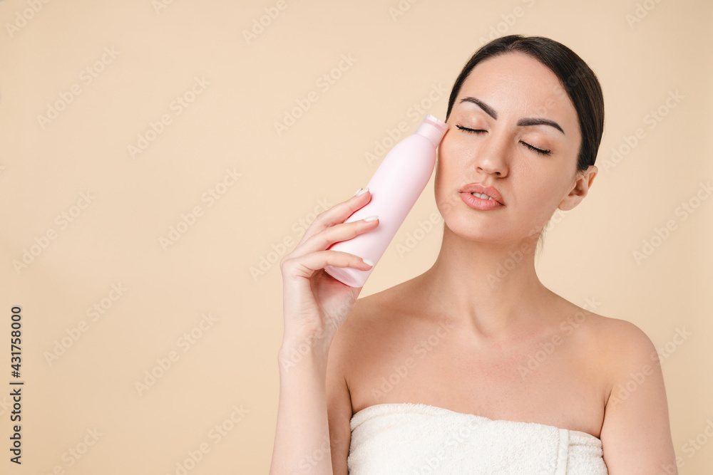 Charming young caucasian woman in bath spa towel ready for taking a shower with shower softing gel isolated over beige background. Pampering and spa effect, skin and body care concept