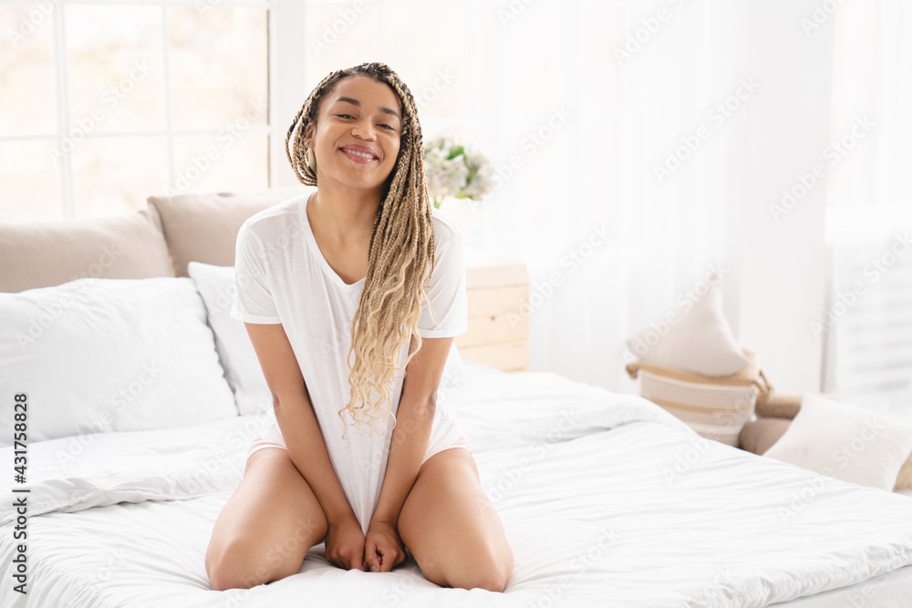 Light image of an african-american young woman girl sitting on comfy bed in white t-shirt, bedding, bedtime, early morning. Let`s start a new day and stay at home Light image of an african-american young woman girl sitting on comfy bed in white t-shirt, bedding, bedtime, early morning. Let`s start a new day and stay at home