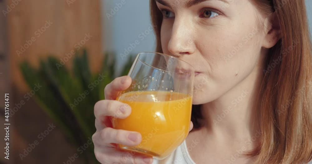 Close up view of the ginger adorable girl drinking healthy orange juice from the glass and looking away with pleasure while enjoying of her morning. Calm girl spending her weekend at home Close up view of the ginger adorable girl drinking healthy orange juice from the glass and looking away with pleasure while enjoying of her morning. Calm girl spending her weekend at home