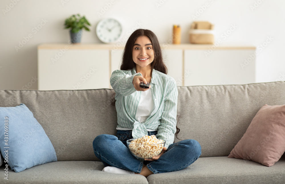 Full length of young Indian woman with remote control watching TV and eating popcorn on sofa at home