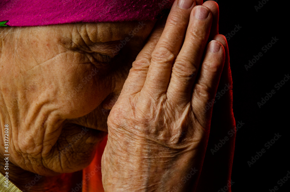 Fototapeta premium An old woman asks for help. An old grandmother in prayer. Grandmother prays on a dark background. Seniors concept. A portrait photo of an old grandmother. A portrait photo of a praying old woman.
