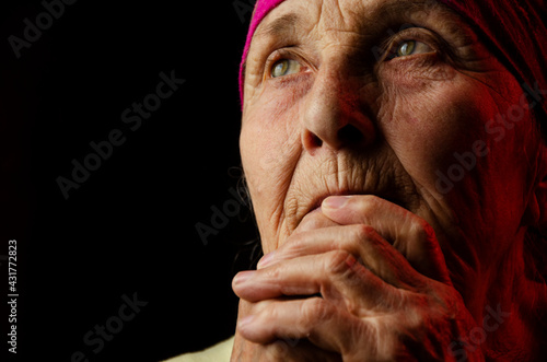 Portrait photo of an old grandmother on a dark background. The wrinkled face of an old woman. Babshuka looks in upward with hope. Pensive old woman. The face of an old grandmother close up.