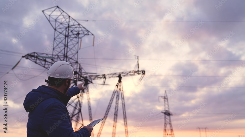 Silhouette of engineer standing on field with electricity towers. Electrical engineer with high voltage electricity pylon at sunset background. Power workers at work concept