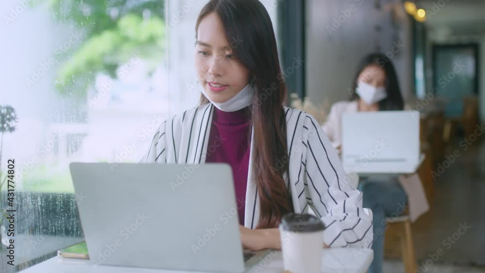 Asian attractive beautiful woman with face mask sitting at table near window at the cafe working using laptop while enjoying her coffee and dessert coffee break Asian attractive beautiful woman with face mask sitting at table near window at the cafe working using laptop while enjoying her coffee and dessert coffee break