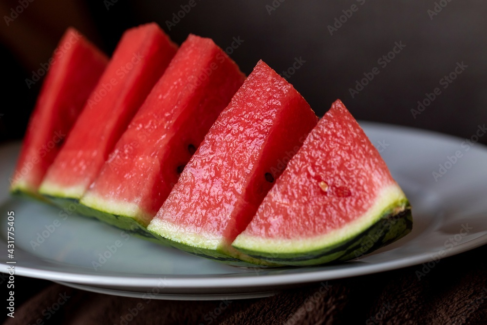 A close up portrait of a sliced up quarter of a watermelon. The fruit is red in the middle with black seeds and has a green striped skin and is lying on a white plate, the background is dark. A close up portrait of a sliced up quarter of a watermelon. The fruit is red in the middle with black seeds and has a green striped skin and is lying on a white plate, the background is dark.