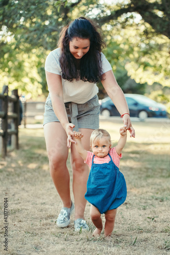 First steps of baby. Cute baby girl learning to walk and holding her mother hand. Mom helping toddler child daughter to go in park outdoor on summer day. Care, assistance and support.