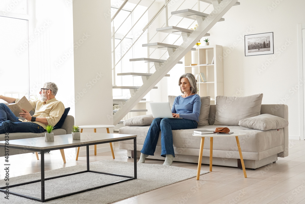 Older senior family couple spending time at home in modern living room. Middle aged husband reading book relaxing sitting on couch while mature wife using laptop enjoying casual daily activities. - obrazy, fototapety, plakaty 