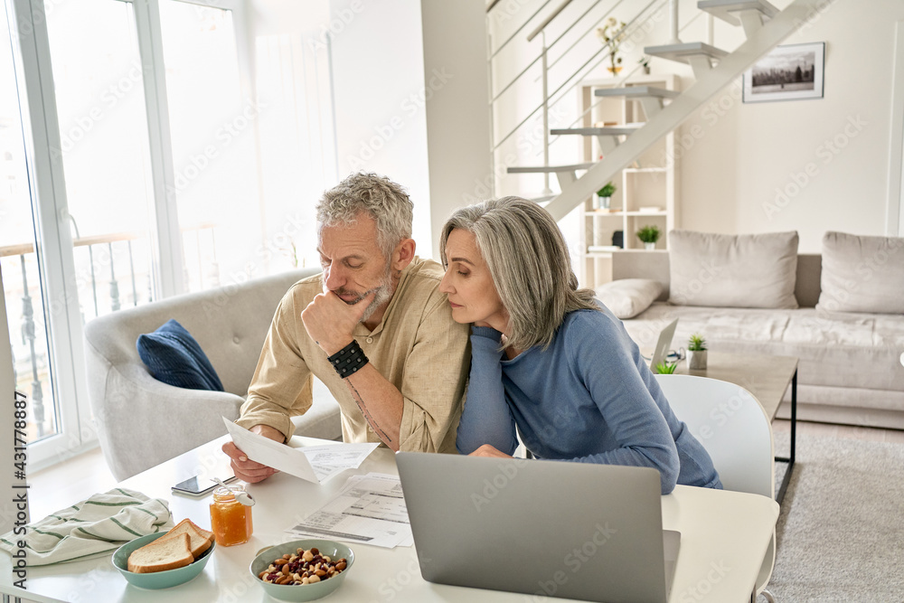 Worried old senior couple checking bank documents at home. Serious mature retired family reading paper bills, calculating taxes, planning retirement finances, household expenses, having money problems Worried old senior couple checking bank documents at home. Serious mature retired family reading paper bills, calculating taxes, planning retirement finances, household expenses, having money problems