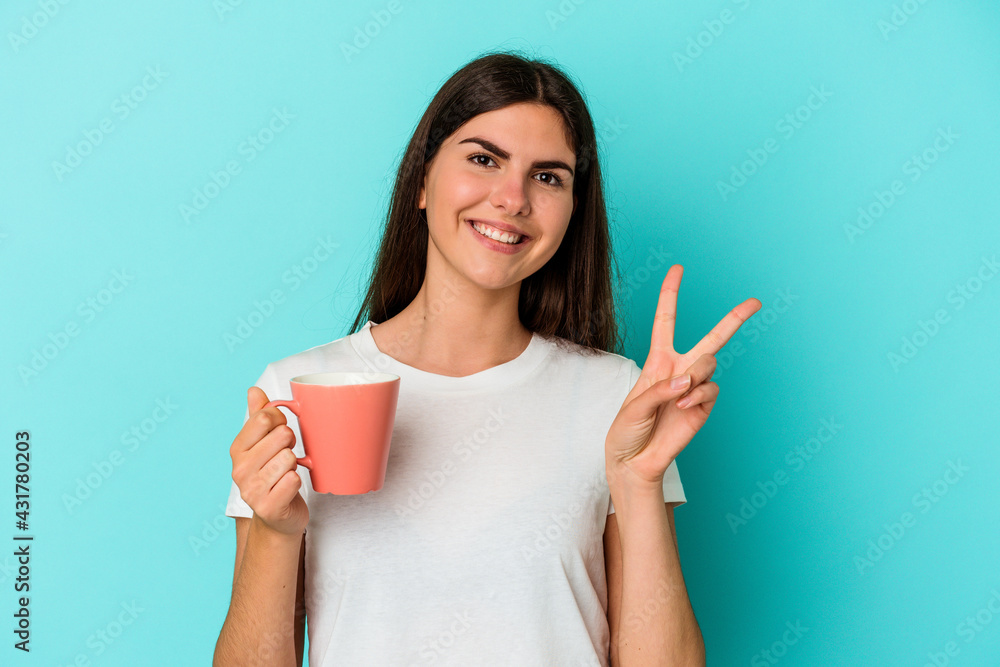 Young caucasian woman holding a mug isolated on blue background showing number two with fingers. Young caucasian woman holding a mug isolated on blue background showing number two with fingers.