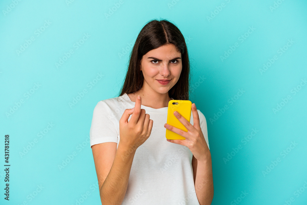 Young caucasian woman holding a mobile phone isolated on blue background pointing with finger at you as if inviting come closer. Young caucasian woman holding a mobile phone isolated on blue background pointing with finger at you as if inviting come closer.