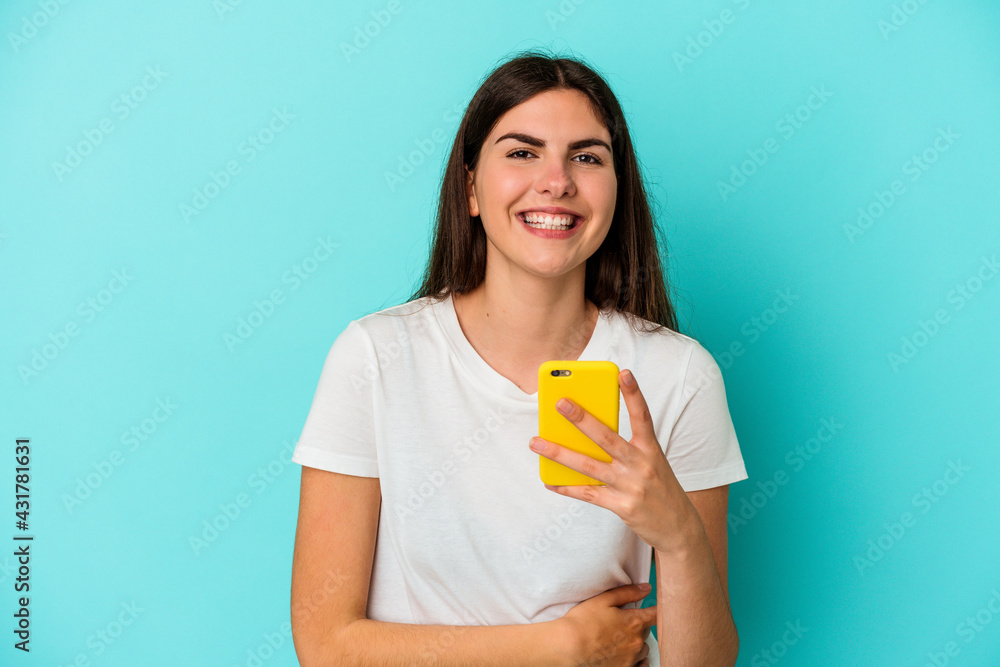 Young caucasian woman holding a mobile phone isolated on blue background laughing and having fun. Young caucasian woman holding a mobile phone isolated on blue background laughing and having fun.