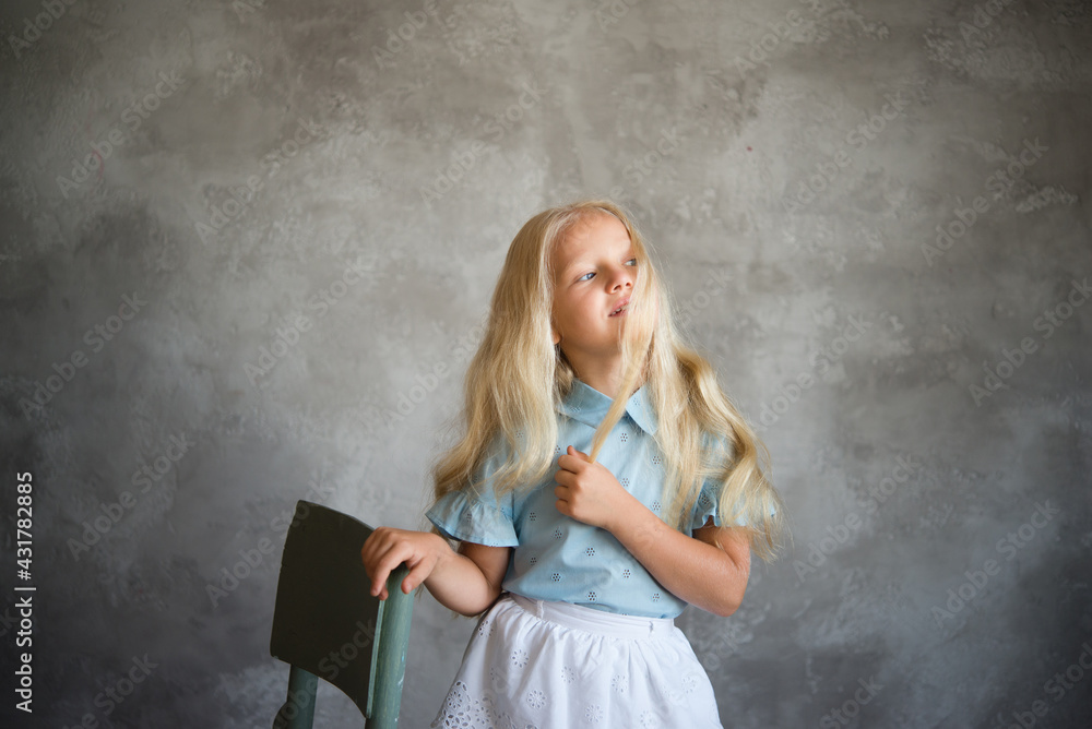 beautiful blonde girl stands near a chair against background of a gray wall. Photo demonstrating the character child - daydreaming, enthusiasm, vulnerability, touching, beauty, stubbornness, cunning beautiful blonde girl stands near a chair against background of a gray wall. Photo demonstrating the character child - daydreaming, enthusiasm, vulnerability, touching, beauty, stubbornness, cunning