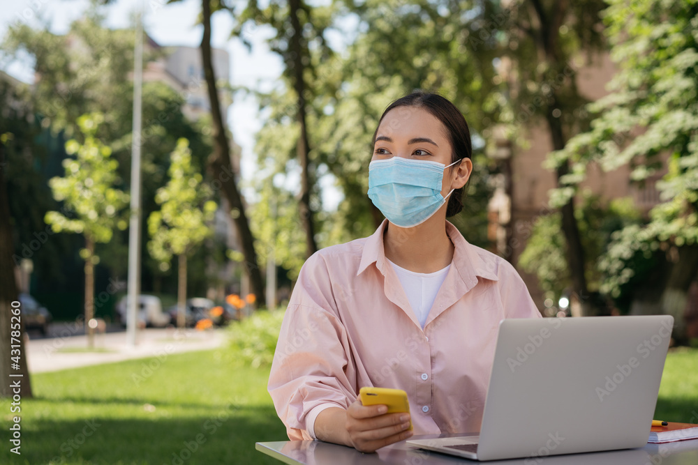 Pensive asian woman wearing medical mask using laptop computer, mobile phone working outdoors. Student studying online, distance learning Pensive asian woman wearing medical mask using laptop computer, mobile phone working outdoors. Student studying online, distance learning