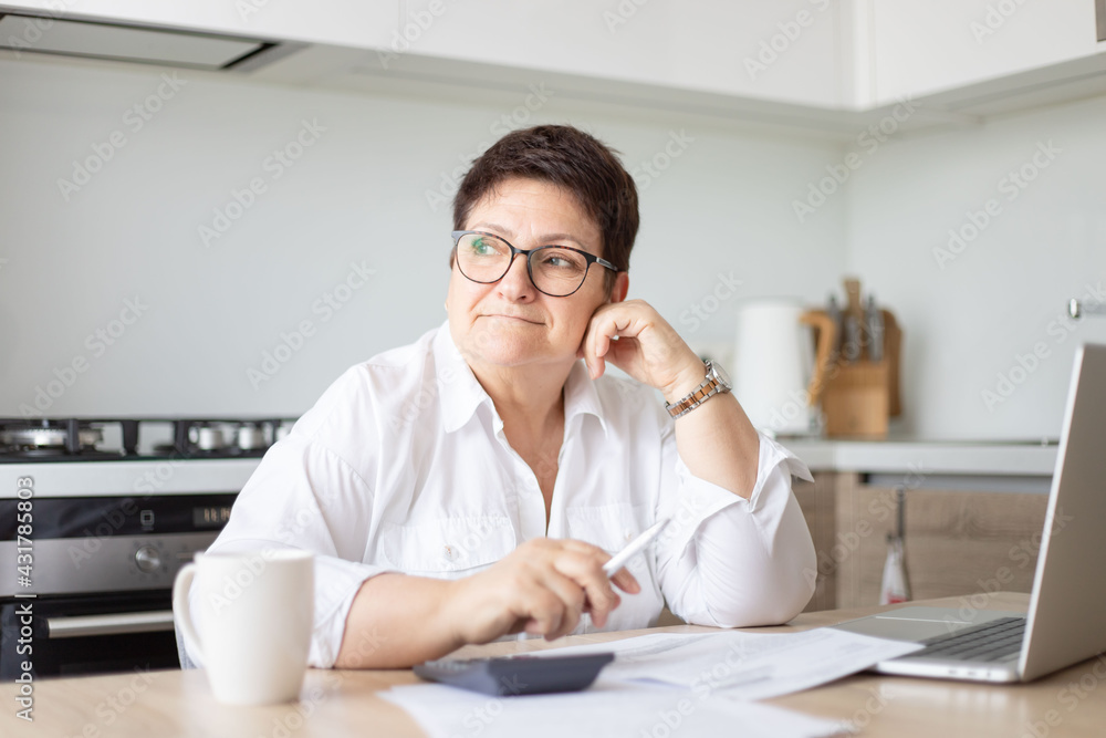 Serious mature Caucasian 60s woman sit at table calculate household finances paying bills on laptop online. Serious mature Caucasian 60s woman sit at table calculate household finances paying bills on laptop online.