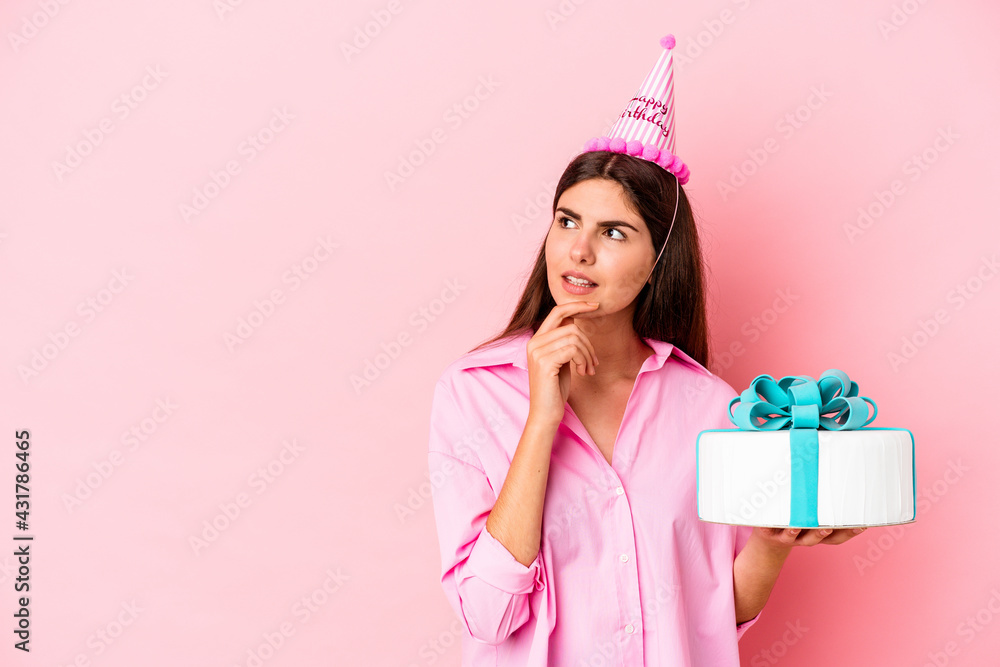 Young caucasian woman holding a cake to celebrate a birthday isolated on pink background looking sideways with doubtful and skeptical expression. Young caucasian woman holding a cake to celebrate a birthday isolated on pink background looking sideways with doubtful and skeptical expression.