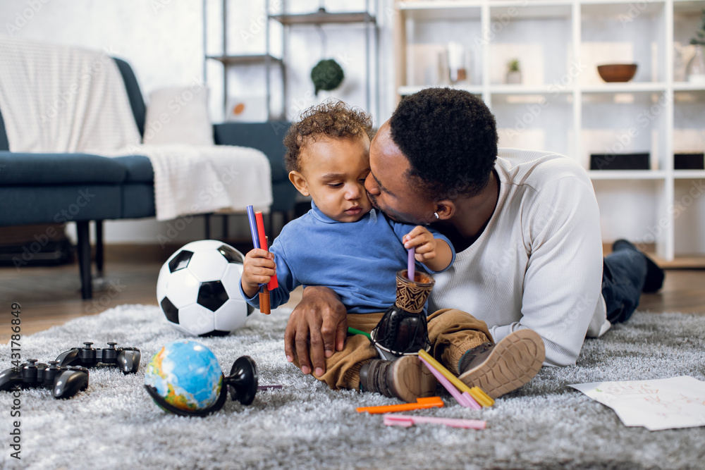 Cute baby boy and african man in casual clothes lying together on soft carpet and playing with different toys. Young father kissing and embracing little son at home. Leisure time.