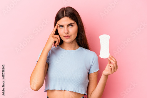 Young caucasian woman holding a compress isolated on pink background pointing temple with finger, thinking, focused on a task.
