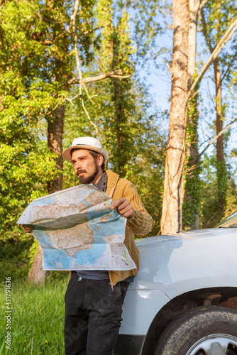 Latino man leaning on his 4wd suv car consulting on a map the route through the forest to follow. lost person with his off-road car. off-track driving. adventure concept in natural environment