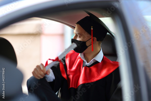 the graduate student in graduation gown and a square cap and a face mask sits in the car before the start of the ceremony