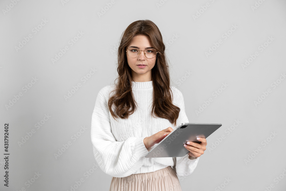 Beautiful girl with glasses working on a tablet, she is seriously looking at the camera. Brunette isolated on gray background. Beautiful girl with glasses working on a tablet, she is seriously looking at the camera. Brunette isolated on gray background.
