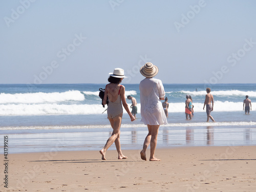 Pareja de dos mujeres mayores paseando en bañador, sombrero y camisa por la playa del Merón en las vacaciones del verano de 2020 en Santander, España. photo