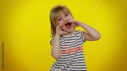 Hey you. Little blonde teen kid child girl in striped t-shirt shouting hello, waving hand, welcoming with hi gesture. Posing isolated on yellow studio background. Young children lifestyle emotions