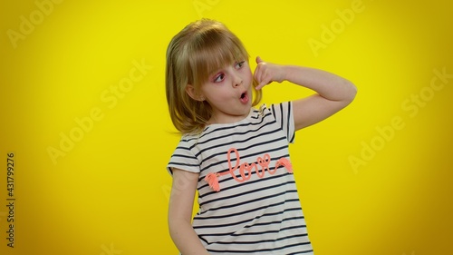 Little cute blonde teen child kid girl in white black striped t-shirt looking at camera doing phone gesture like says hey you call me back. Young children posing isolated on yellow studio background