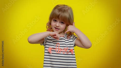 Portrait of funny playful blonde child girl 5-6 years old in striped t-shirt looking at camera doing phone gesture like says hey you call me back on yellow studio wall background. Teen kid children