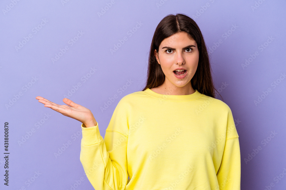 Young caucasian woman isolated on purple background impressed holding copy space on palm. Young caucasian woman isolated on purple background impressed holding copy space on palm.