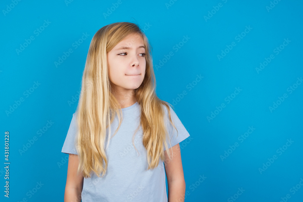 Amazed puzzled beautiful Caucasian little girl wearing white T-shirt over blue background, curves lips and has worried look, sees something awful in front. Amazed puzzled beautiful Caucasian little girl wearing white T-shirt over blue background, curves lips and has worried look, sees something awful in front.