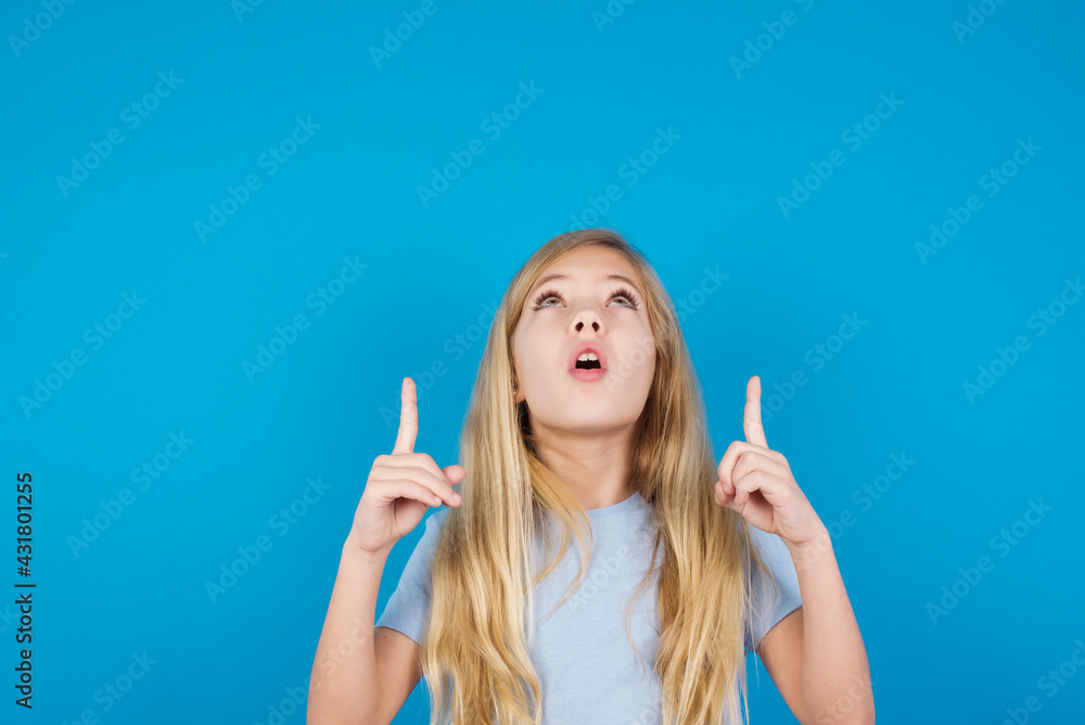 Obraz premium beautiful Caucasian little girl wearing white T-shirt over blue background being amazed and surprised looking and pointing up with fingers showing something strange.