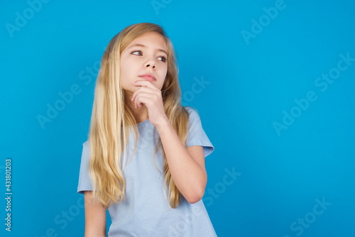 Shot of contemplative thoughtful beautiful Caucasian little girl wearing white T-shirt over blue background keeps hand under chin, looks thoughtfully upwards.
