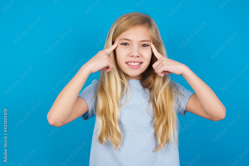 beautiful Caucasian little girl wearing blue T-shirt over blue background concentrating hard on an idea with a serious look, thinking with both index fingers pointing to forehead.