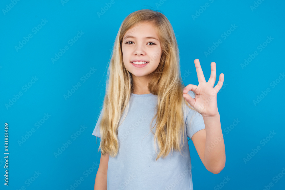 Glad attractive beautiful Caucasian little girl wearing blue T-shirt over blue background shows ok sign with hand as expresses approval, has cheerful expression, being optimistic. Glad attractive beautiful Caucasian little girl wearing blue T-shirt over blue background shows ok sign with hand as expresses approval, has cheerful expression, being optimistic.