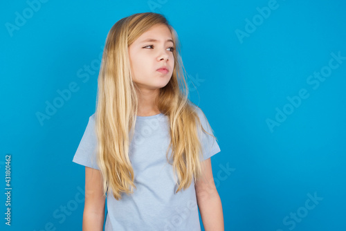 beautiful Caucasian little girl wearing blue T-shirt over blue background  looks pensively aside  plans actions after university  imagines what to do Thinks over about new project.