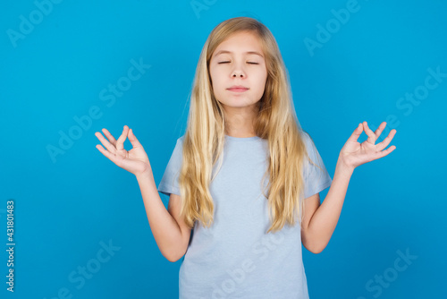 beautiful Caucasian little girl wearing blue T-shirt over blue background doing yoga, keeping eyes closed, holding fingers in mudra gesture. Meditation, religion and spiritual practices. © Jihan