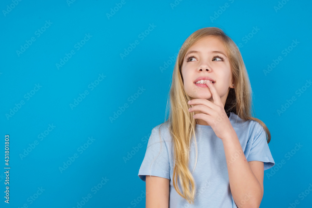 beautiful Caucasian little girl wearing blue T-shirt over blue background with thoughtful expression, looks to the camera, keeps hand near face, bitting a finger thinks about something pleasant. beautiful Caucasian little girl wearing blue T-shirt over blue background with thoughtful expression, looks to the camera, keeps hand near face, bitting a finger thinks about something pleasant.