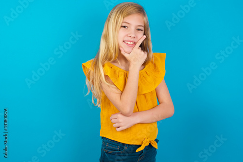 Caucasian kid girl wearing yellow T-shirt against blue wall looking confident at the camera smiling with crossed arms and hand raised on chin. Thinking positive.