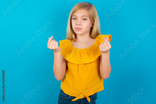 Caucasian kid girl wearing yellow T-shirt against blue wall doing money gesture with hands, asking for salary payment, millionaire business