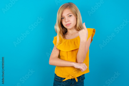 Caucasian kid girl wearing yellow T-shirt against blue wall Beckoning come here gesture with hand inviting welcoming happy and smiling