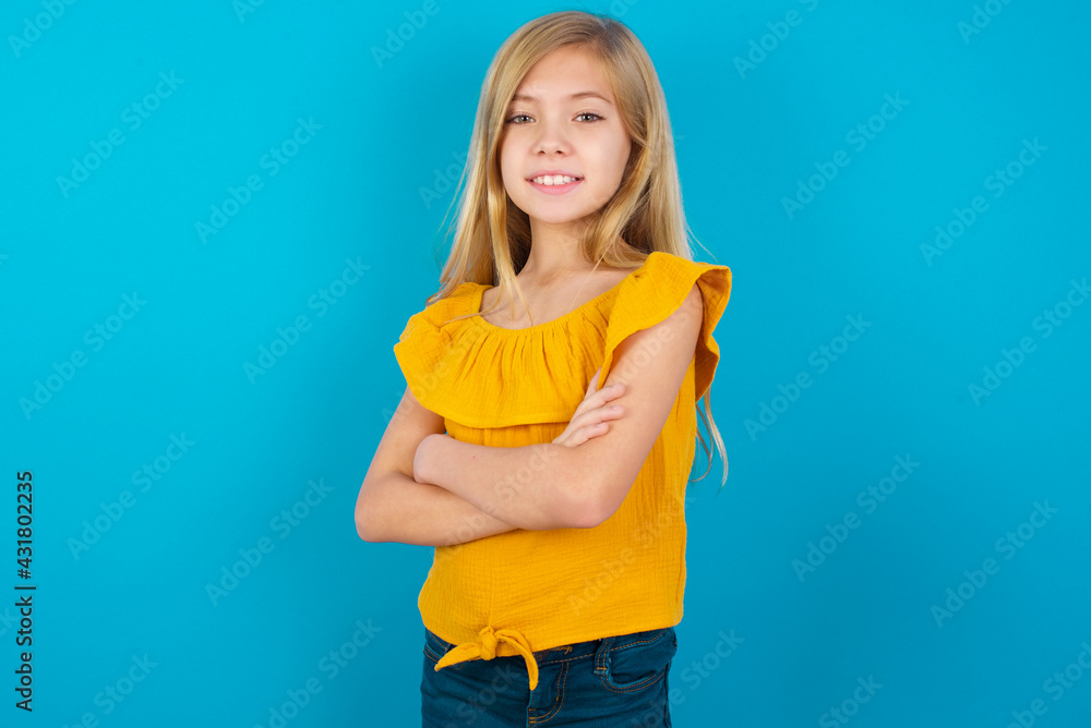 Caucasian kid girl wearing yellow T-shirt against blue wall happy face smiling with crossed arms looking at the camera. Positive person. Caucasian kid girl wearing yellow T-shirt against blue wall happy face smiling with crossed arms looking at the camera. Positive person.