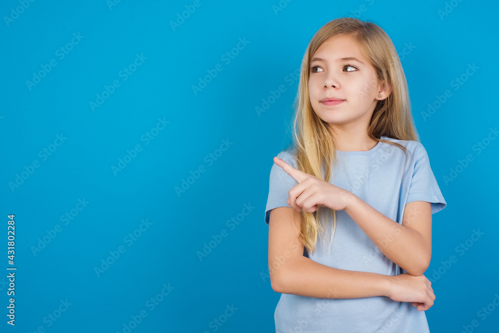 Portrait of beautiful Caucasian little girl wearing blue T-shirt over blue background posing on camera with tricky look, presenting product with index finger. Advertisement concept. Portrait of beautiful Caucasian little girl wearing blue T-shirt over blue background posing on camera with tricky look, presenting product with index finger. Advertisement concept.