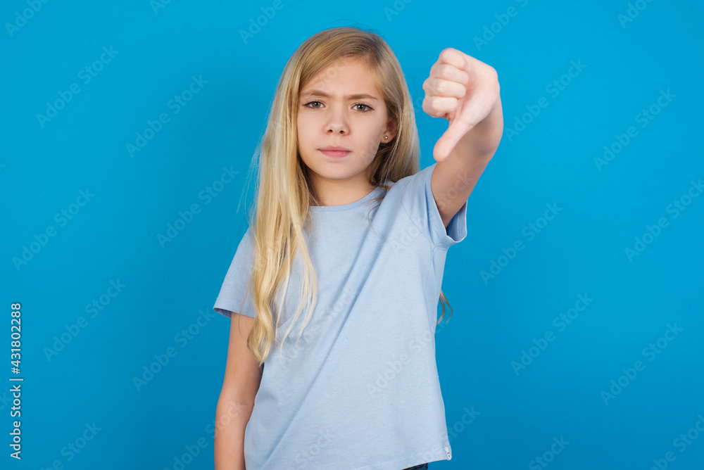 Discontent beautiful Caucasian little girl wearing white T-shirt over blue background shows disapproval sign, keeps thumb down, expresses dislike, frowns face in discontent. Negative feelings. Discontent beautiful Caucasian little girl wearing white T-shirt over blue background shows disapproval sign, keeps thumb down, expresses dislike, frowns face in discontent. Negative feelings.