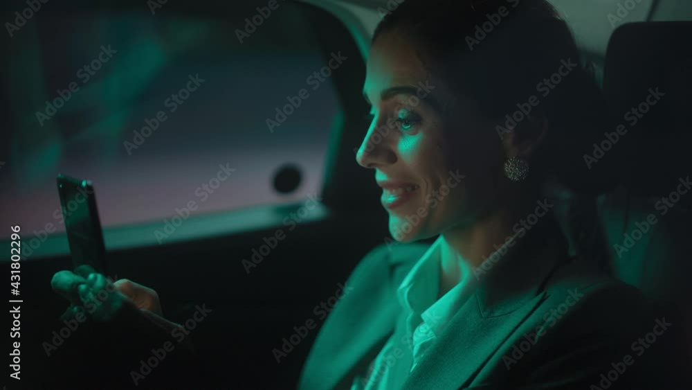 Portrait of Successful Businesswoman Commuting from Office in a Backseat of Her Car at Night. Entrepreneur Using Smartphone while in Transfer Taxi in Urban City Street with Working Neon Signs. Portrait of Successful Businesswoman Commuting from Office in a Backseat of Her Car at Night. Entrepreneur Using Smartphone while in Transfer Taxi in Urban City Street with Working Neon Signs.