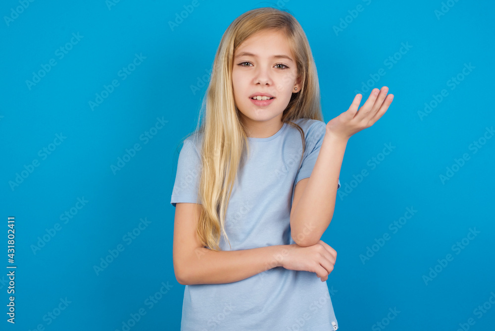 Fototapeta premium Studio shot of frustrated beautiful Caucasian little girl wearing blue T-shirt over blue background gesturing with raised palm, frowning, being displeased and confused with dumb question.