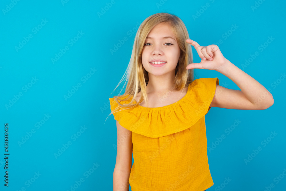 Caucasian kid girl wearing yellow T-shirt against blue wall smiling and confident gesturing with hand doing small size sign with fingers looking and the camera. Measure concept Caucasian kid girl wearing yellow T-shirt against blue wall smiling and confident gesturing with hand doing small size sign with fingers looking and the camera. Measure concept